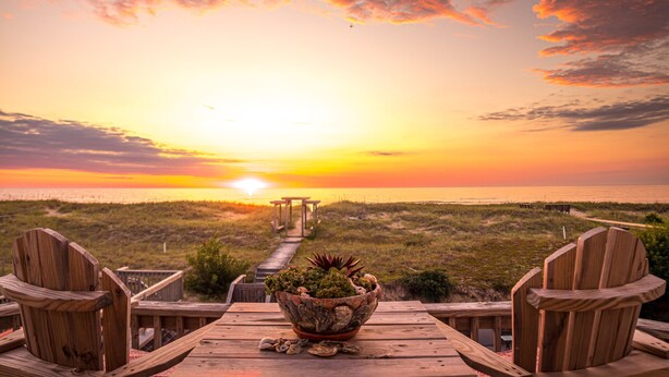 A wooden deck with a table and chairs, a bowl of plants, and a sunset view over the beach.