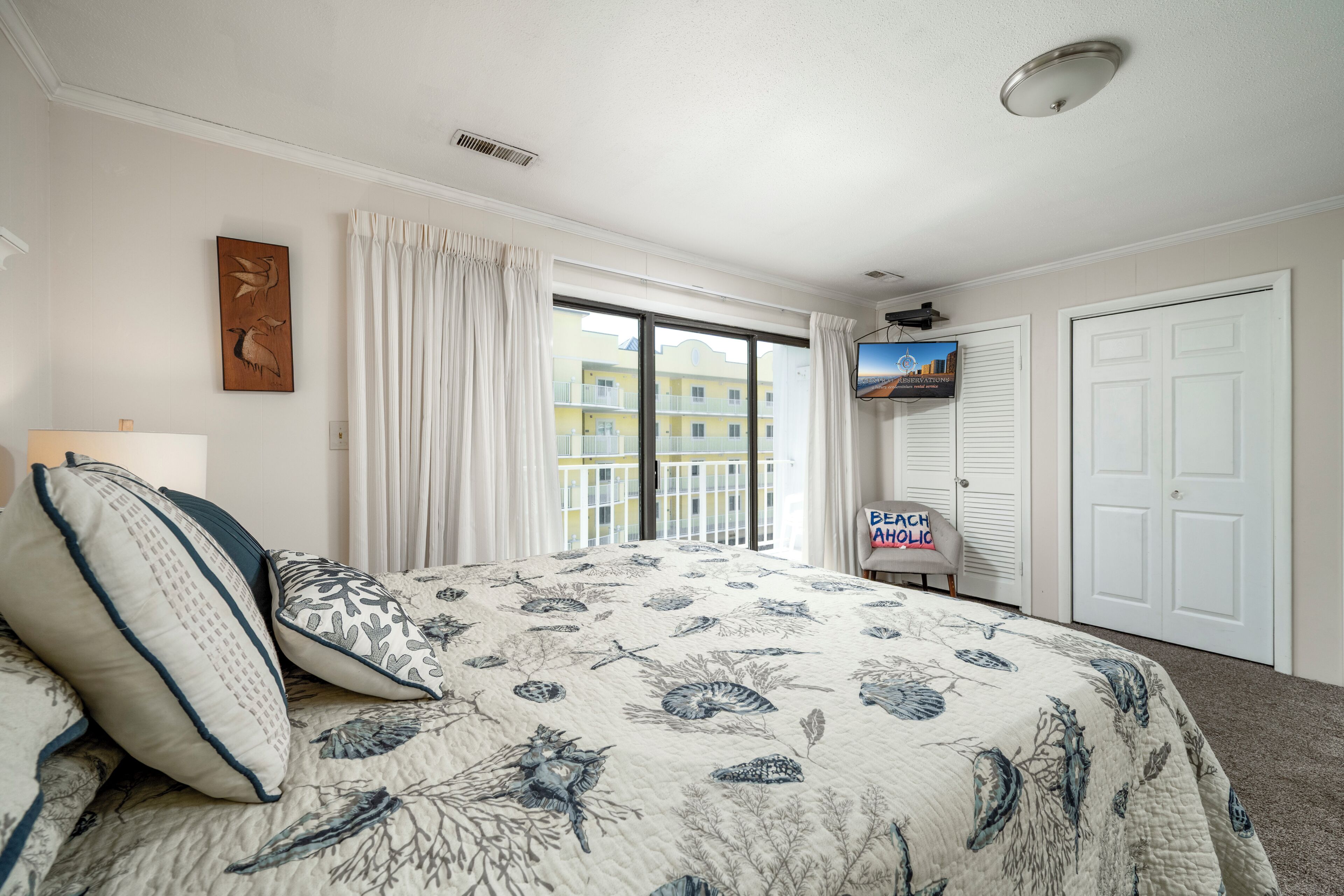 Ein Schlafzimmer mit einem großen Bett, einem Fernseher und Blick auf Gebäude. Ein Schild trägt die Aufschrift 'Beach Aloha'.