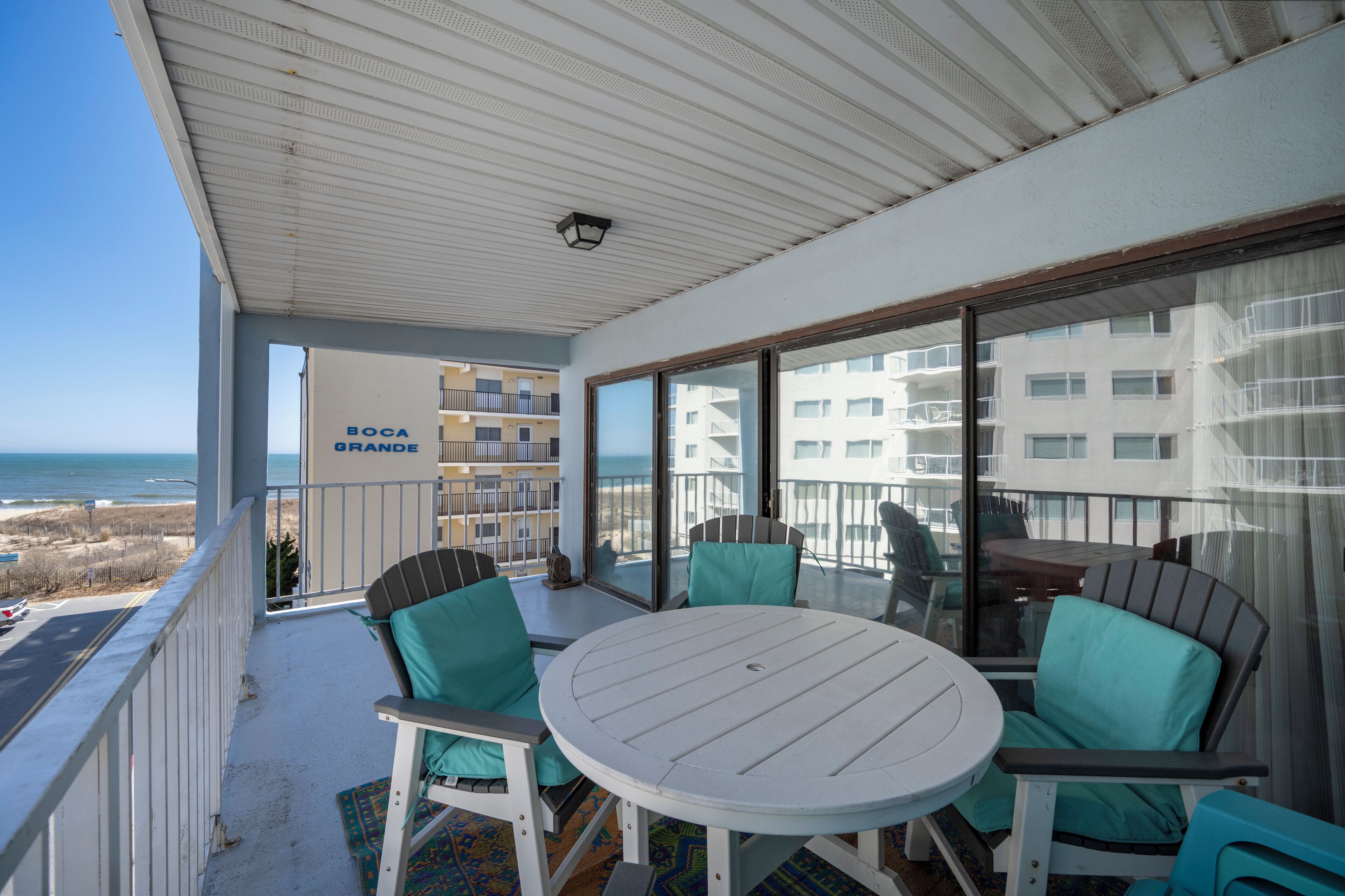 Un balcón con una mesa redonda y sillas, con vista a una playa y un edificio con la leyenda "BOCA GRANDE".