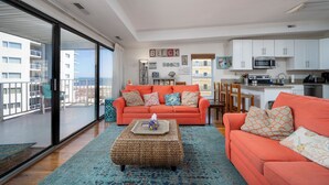 Un amplio salón con un gran ventanal, un sofá naranja brillante, un reposapiés tejido y una cocina al fondo.