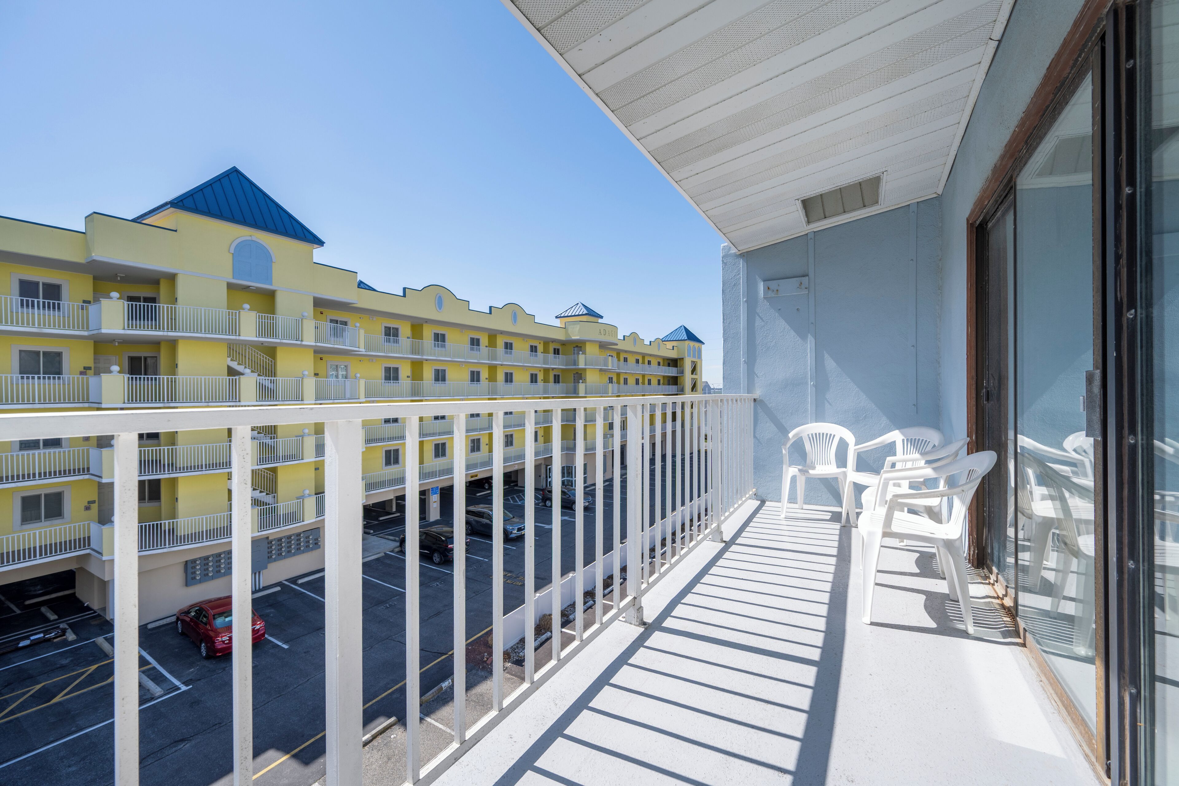 A balcony with white chairs, a view of a yellow building with blue roofs, and a parking area with cars.