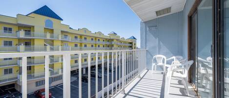 A balcony with white chairs, a view of a yellow building with blue roofs, and a parking area with cars.