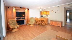 Un salon avec un plancher en bois, un téléviseur, une cheminée, un coin repas avec des chaises et une table, et une cuisine à l’arrière-plan.