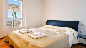 A hotel room with a bed, a window with a view of a cactus and a chair outside, and a small bedside table.