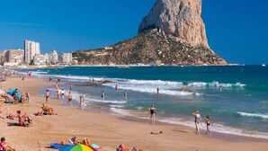 A beach with a prominent rocky hill, clear blue sky, and a cityscape in the background.