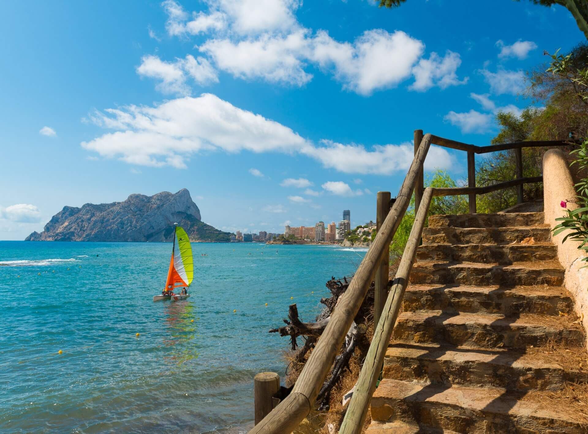 A coastal area with a wooden staircase leading down to the beach, a sailboat with a colorful sail, and a mountain in the background.