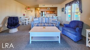 A living room with a striped sofa, a blue armchair, a white coffee table, and a ceiling fan. - Surf Condos 222 - Community Pool & Beach Bliss! (Surf City)