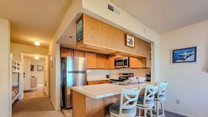 A compact kitchen with wooden cabinets, a stainless steel refrigerator, a microwave, and a coffee maker. There are three bar stools and a small island with a sink. - Surf Condos 222 - Community Pool & Beach Bliss! (Surf City)