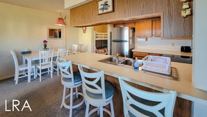 A modern kitchen with wooden cabinets, a stainless steel refrigerator, and a dining area with white chairs and a table. - Surf Condos 222 - Community Pool & Beach Bliss! (Surf City)