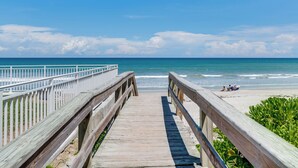 Une promenade en bois mène à une plage d’où l’on a une vue dégagée sur l’océan, et où l’on aperçoit quelques personnes au loin.