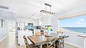 Une cuisine moderne avec des meubles blancs, un îlot central et un coin repas avec des chaises en bois. Une grande fenêtre offre une vue sur la plage.