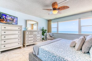 A bedroom with a bed, a dresser, a chair, a ceiling fan, a large window, and a TV.