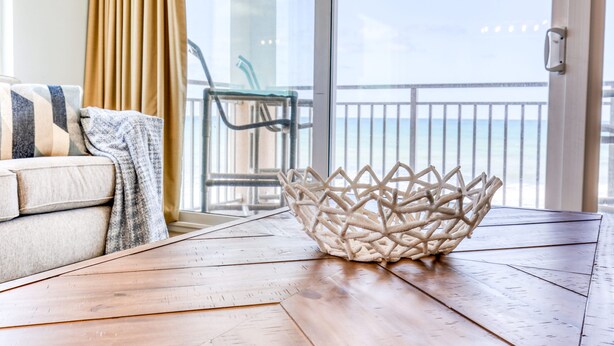 Une table en bois avec un panier tressé, un canapé recouvert d’un plaid et une vue sur la mer par une porte vitrée.