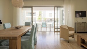 En moderne stue med et træspisebord, lyseblå polstrede stole, en hængende lysarmatur i flet, en beige lænestol og et fjernsyn på et træstel.