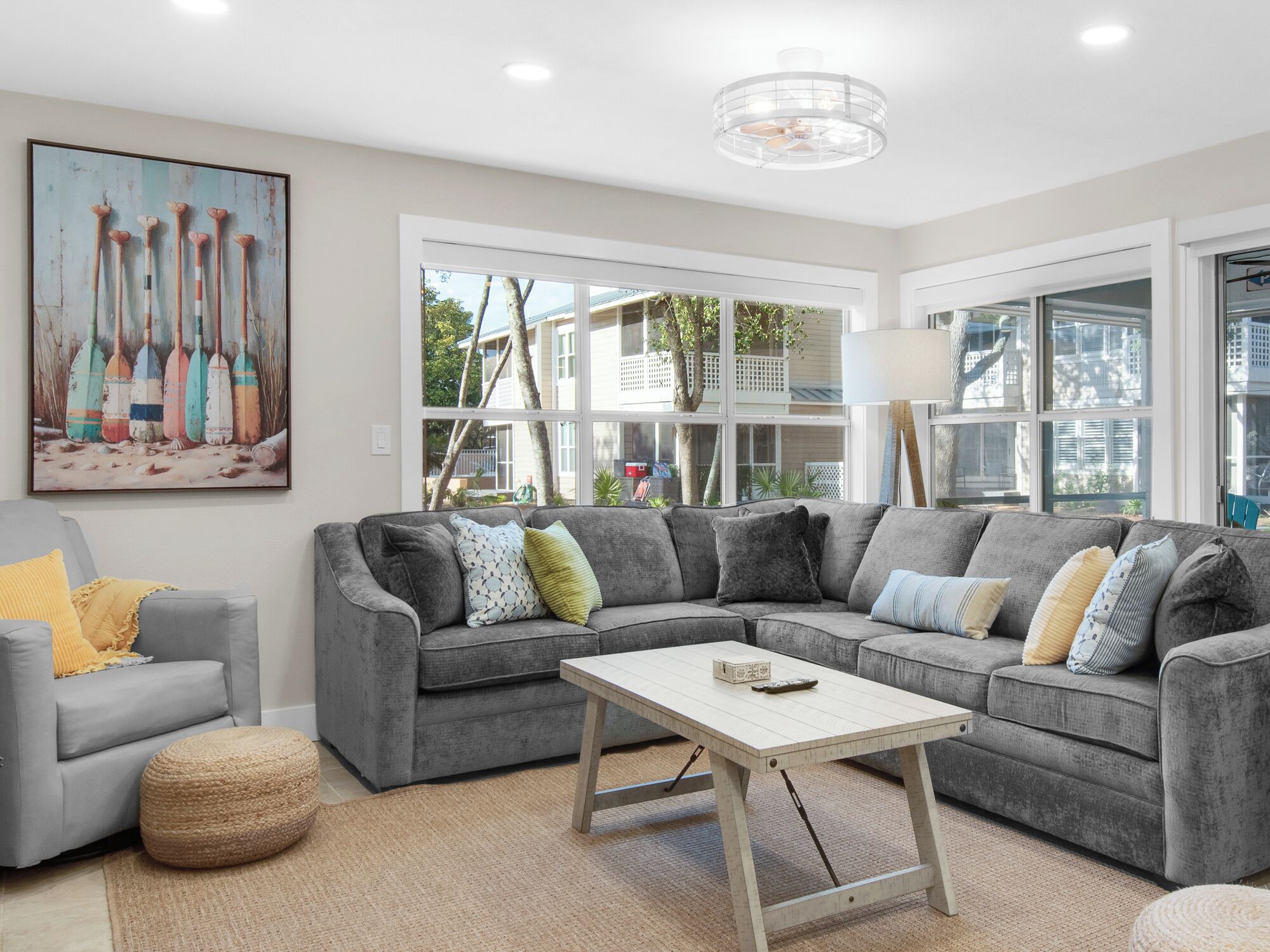 Un salon moderne avec un canapé d’angle gris, un petit fauteuil gris et une table basse blanche. Une grande fenêtre offre une vue sur une maison et des arbres à l’extérieur.