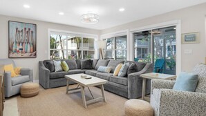 Ein modernes Wohnzimmer mit einem grauen Ecksofa, einem Couchtisch und einem kleinen Beistelltisch. An der Wand hängt ein Gemälde, ein Fenster bietet Blick ins Freie und ein Schild mit der Aufschrift 'GULF COAST'.