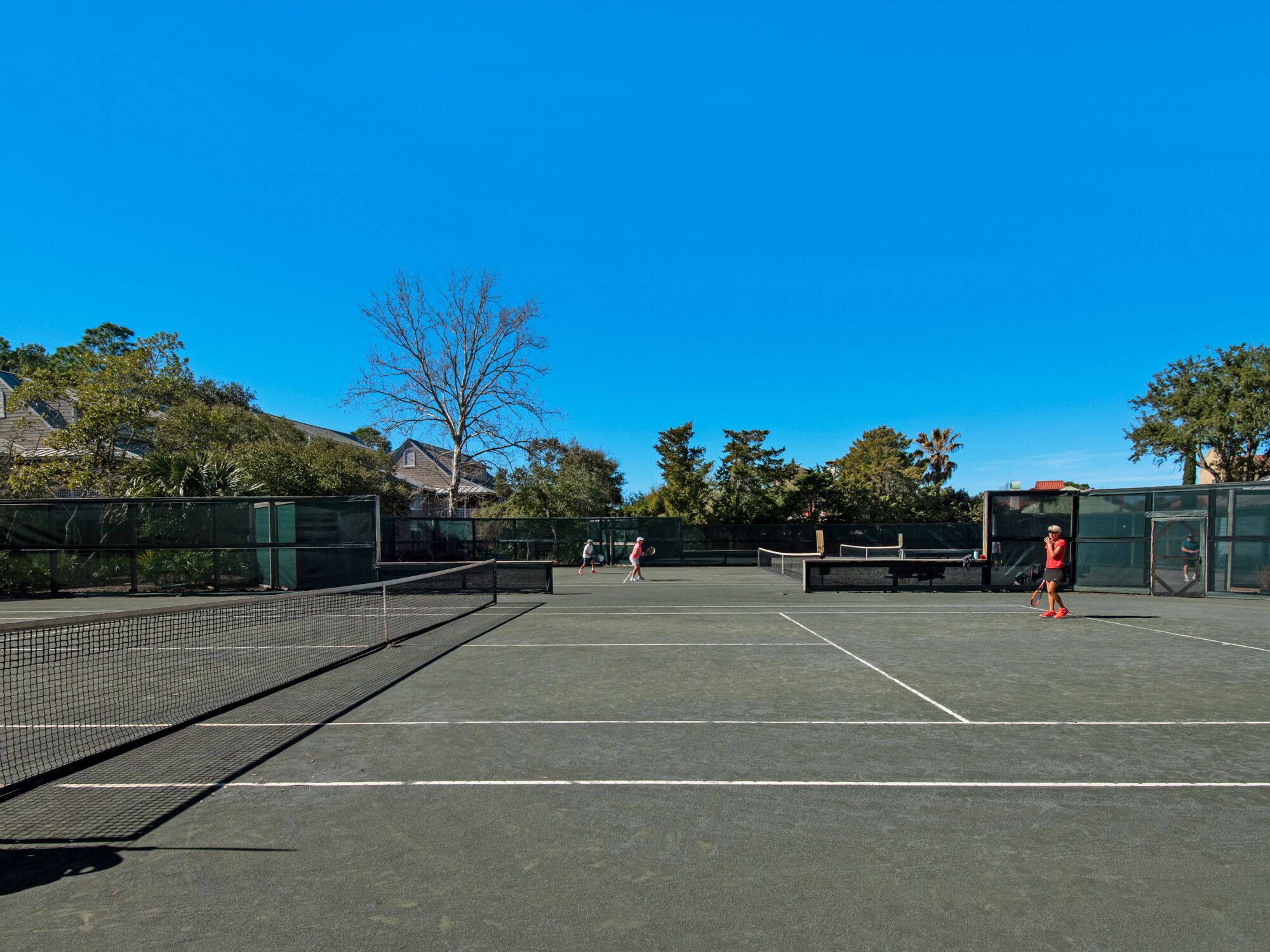 An outdoor tennis court with a net, surrounded by trees and a building in the background.