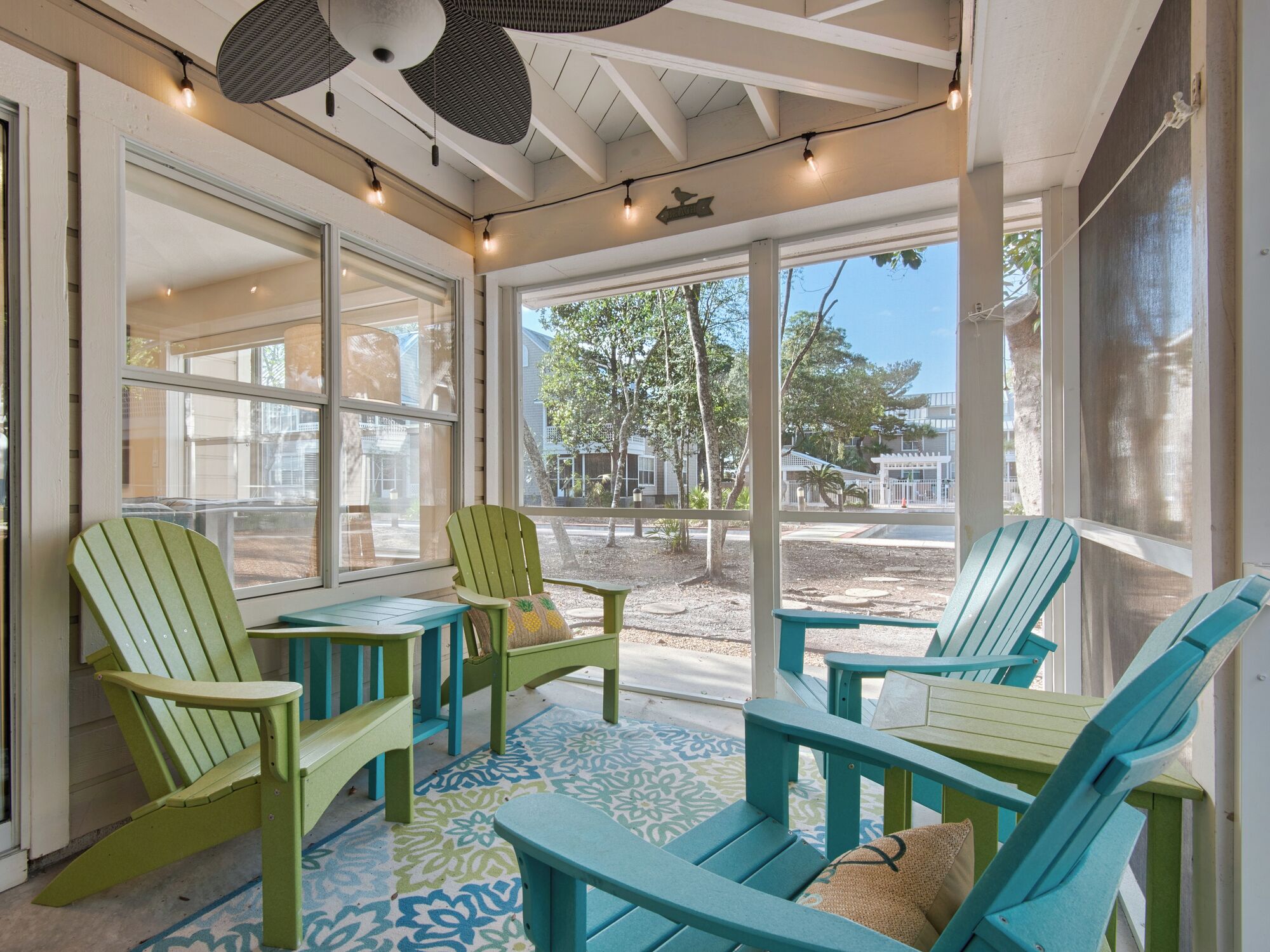 A covered outdoor seating area with green and blue Adirondack chairs, a patterned floor, and large windows offering a view of the outdoors.