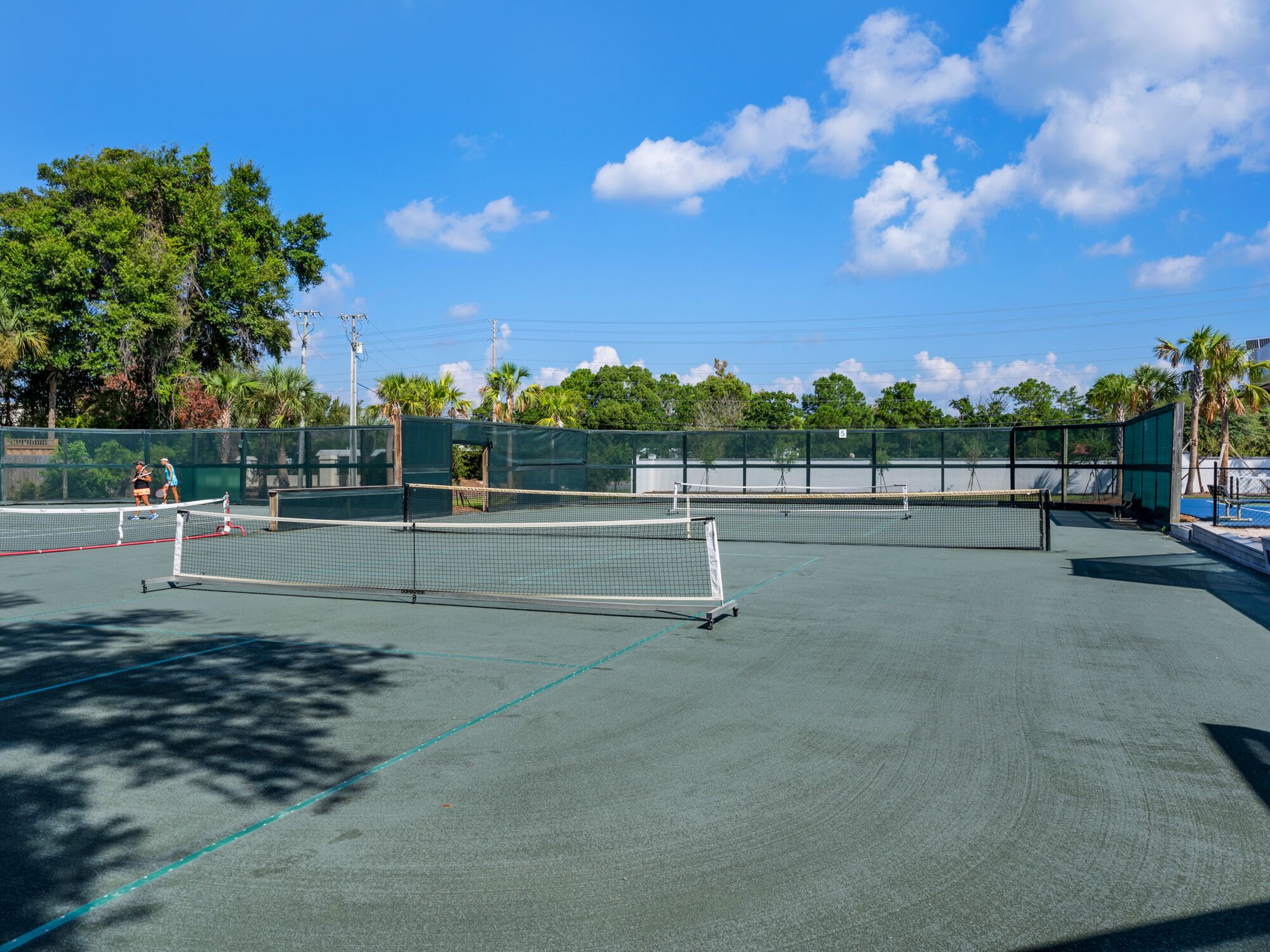 An outdoor tennis court with multiple courts, surrounded by trees and a clear sky.