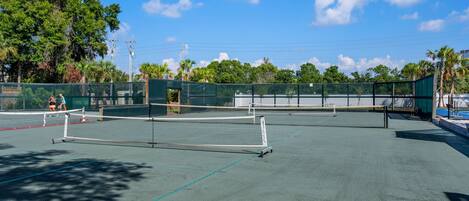An outdoor tennis court with multiple courts, surrounded by trees and a clear sky.