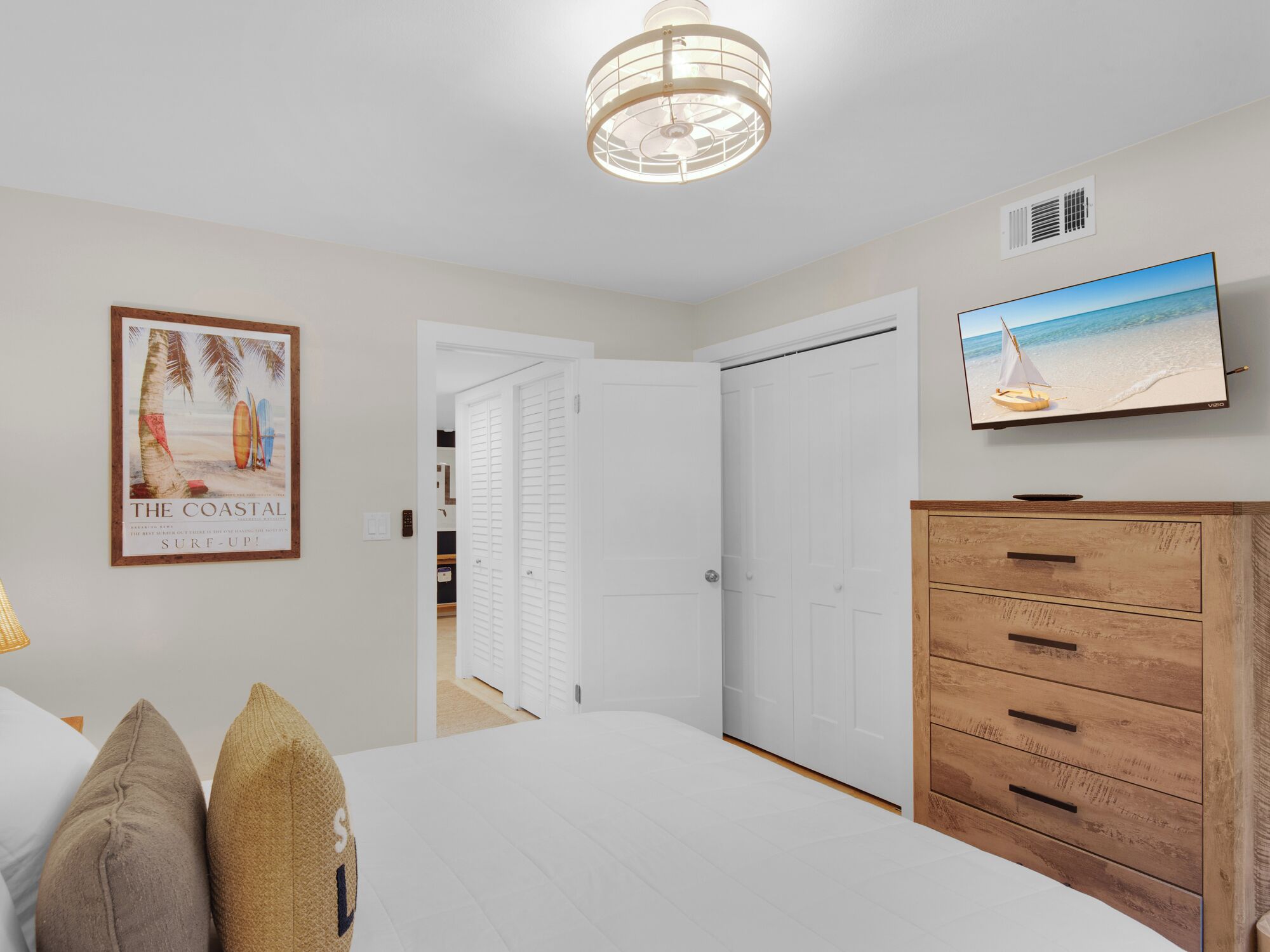 A bedroom with a bed, a wooden dresser, a television on the wall, a framed picture of a beach, and a door leading to another room.