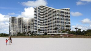 A large, multi-story hotel with a curved facade, located near a beach with palm trees and a sandy area. - Great Beach Views, Direct Access, Pickleball, New Kitchen & Heated Pool! Tower 3 (Marco Island)