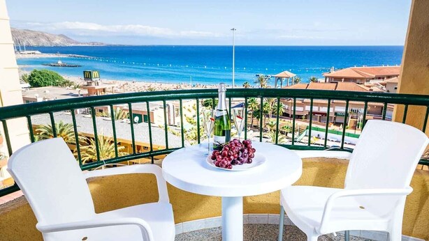 En balkon med to hvide plastikstole og et lille rundt bord med en flaske og druer. Fra balkonen er der udsigt til en strand med bygninger og en klar blå himmel.
