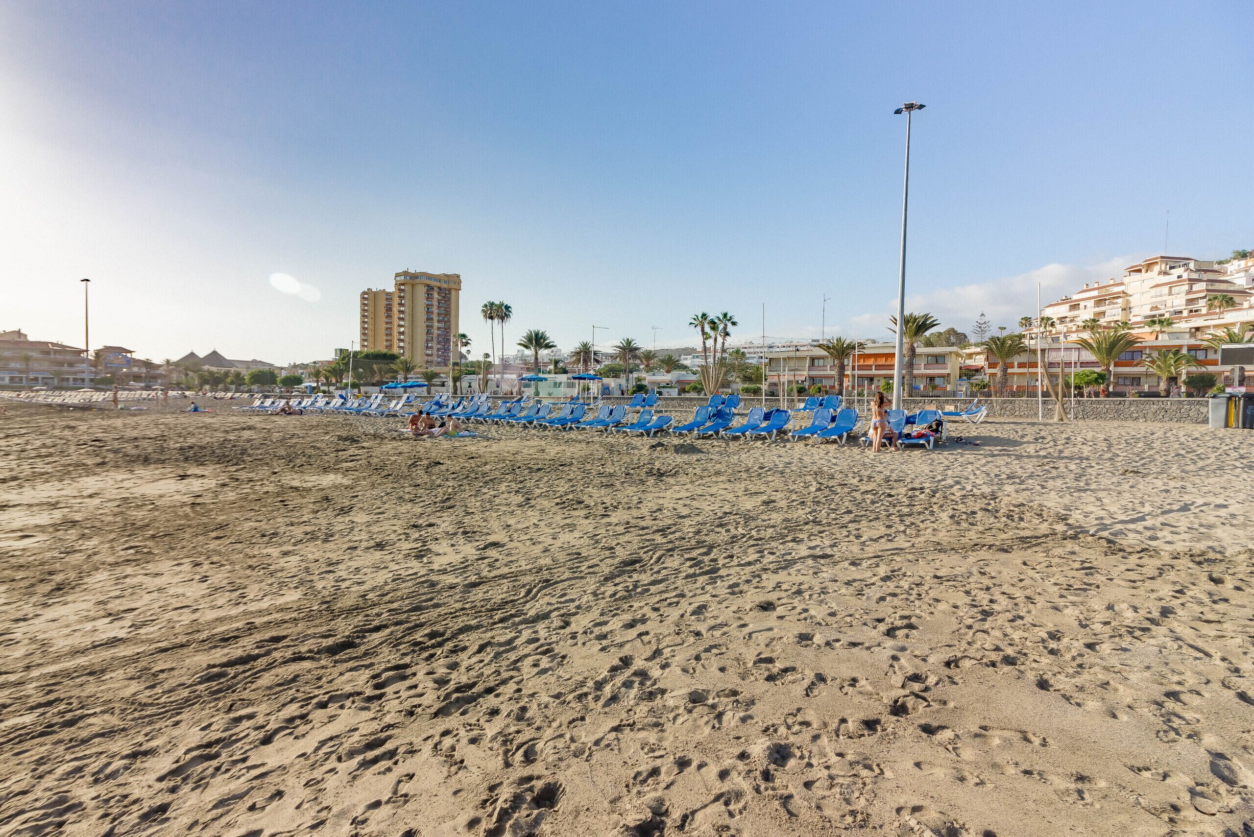A sandy beach with rows of blue beach chairs and umbrellas, a tall building in the background, and a clear sky.