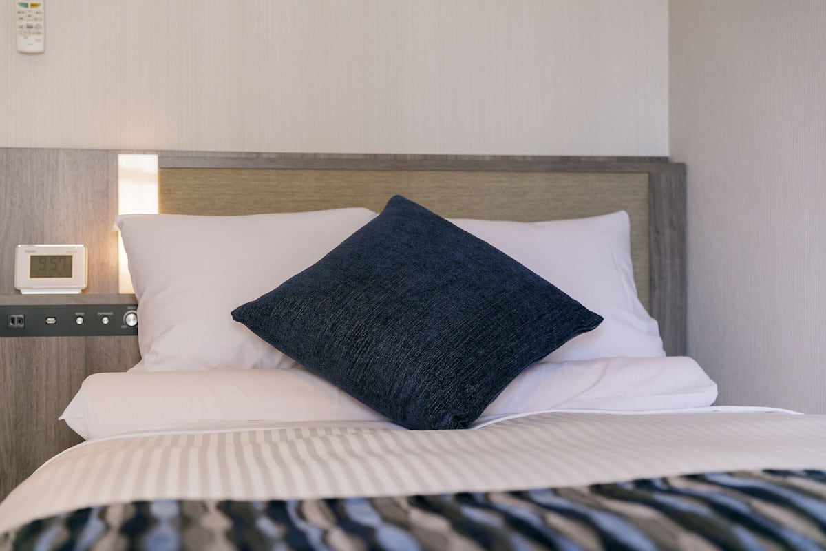 Een netjes opgemaakt bed met witte lakens en een donkerblauwe kussen, een klein nachtkastje met een digitale klok, en een houten hoofdbord.