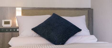 A neatly made bed with white linens and a dark blue pillow, a small bedside table with a digital clock, and a wooden headboard.