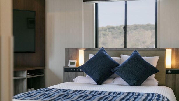 A hotel room with a bed, two bedside lamps, a nightstand, a TV, and a window with a view of trees. - H​OTEL MYSTAYS Kagoshima Tenmonkan Annex (Kagoshima)
