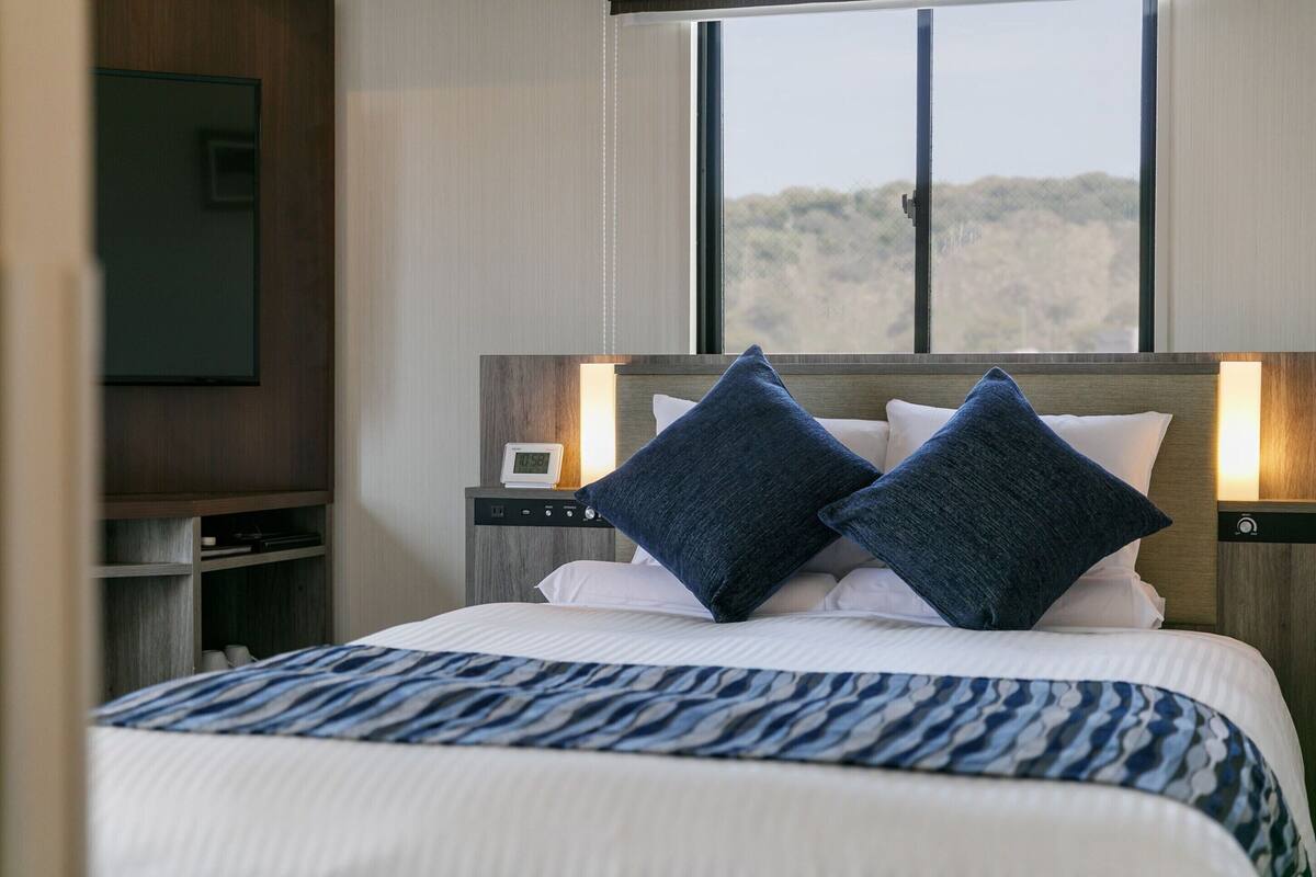 Hotelkamer met een bed, twee bedlampjes, een nachtkastje, een televisie en een raam met uitzicht op bomen.