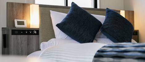 A neatly made bed with two pillows, a bedside table with a digital clock, and a window with blinds.
