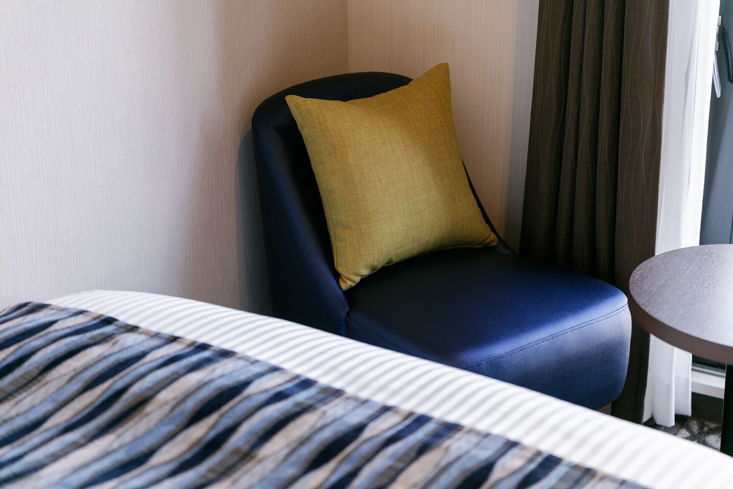 A hotel room with a bed, a blue armchair with a mustard-colored pillow, a round table, and a window with curtains.