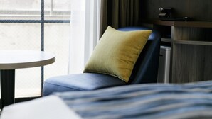 A hotel room with a bed, a chair with a pillow, a small table, a TV, and a window with a view of buildings. - H​OTEL MYSTAYS Kagoshima Tenmonkan Annex (Kagoshima)