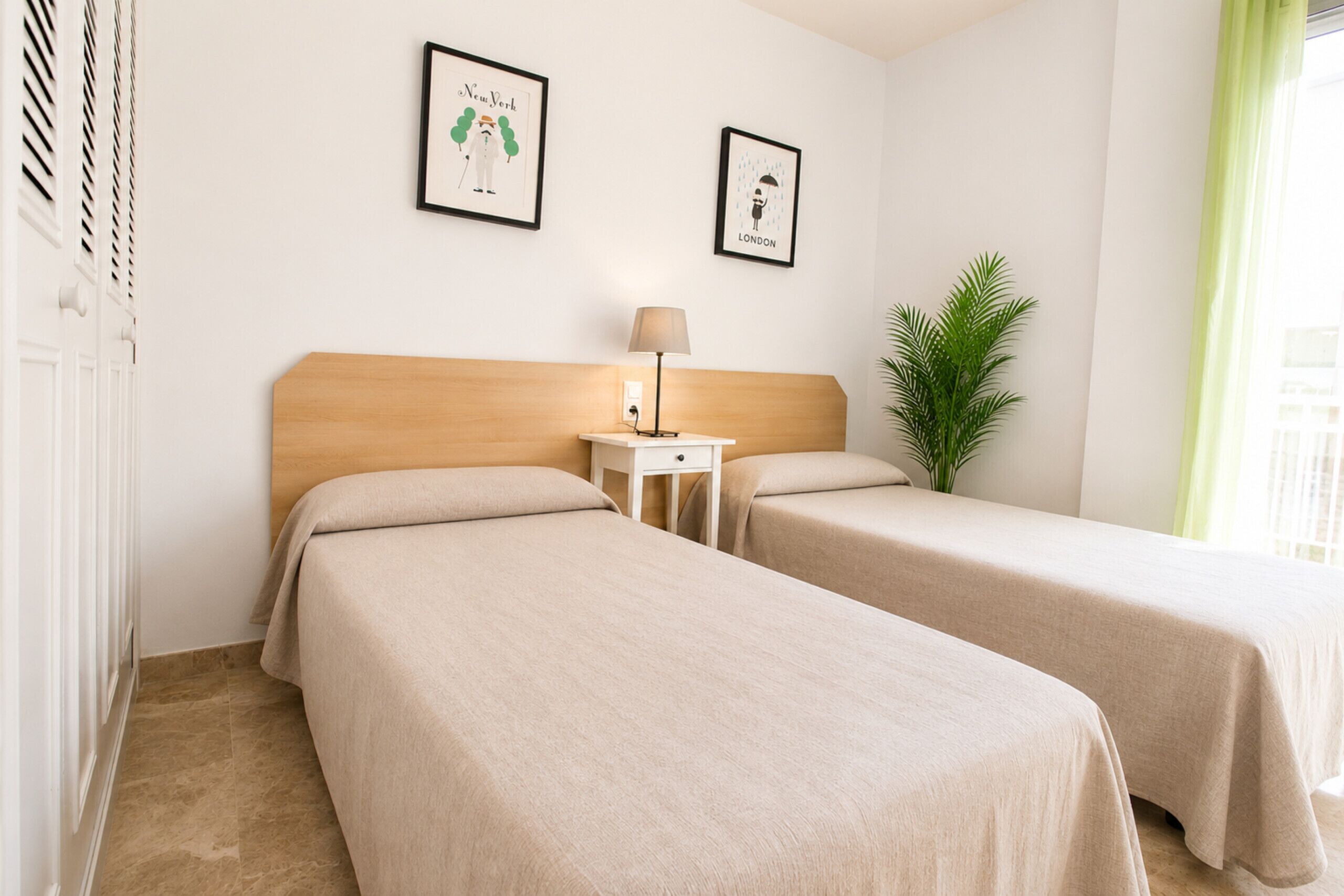 Une chambre d’hôtel avec deux lits, une table de chevet, une lampe, un tableau encadré représentant New York, un tableau encadré représentant Londres et une plante en pot.