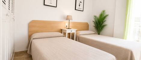 A hotel room with two beds, a nightstand, a lamp, a framed picture of New York, a framed picture of London, and a potted plant.