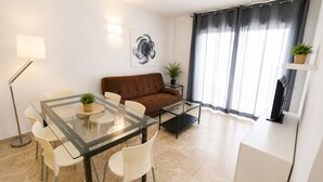 En moderne stue med et glasbord, hvide stole, en brun sofa, et sort sofabord, en potteplante, et indrammet kunstværk og et fjernsyn på et hvidt skab.