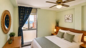 A hotel room with a bed, a ceiling fan, a window with curtains, a wall-mounted mirror, and a flower painting. - DMS Apartment (La Pineda)