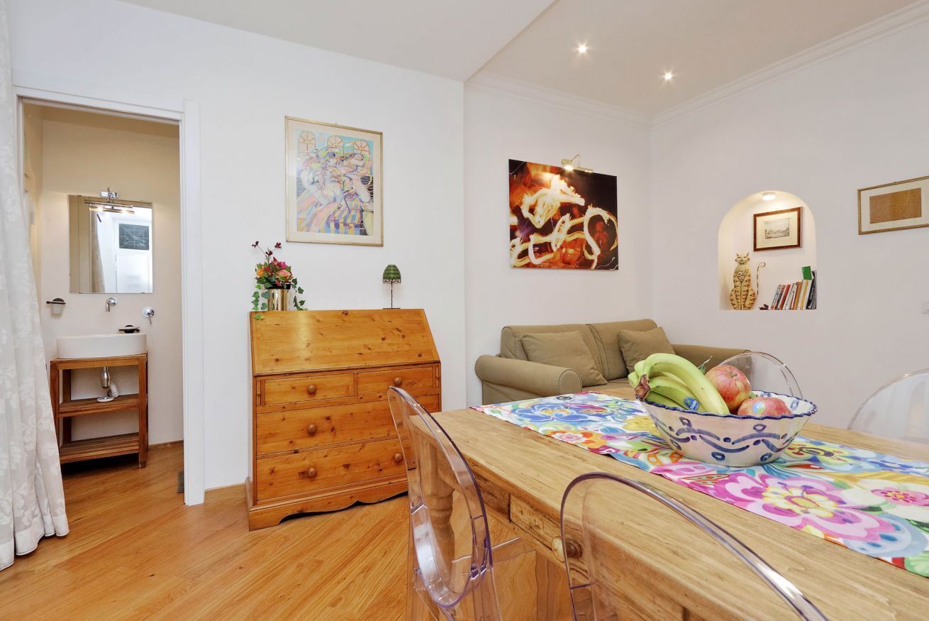 Une pièce comprenant une commode en bois, un canapé, une table à manger recouverte d’une nappe à motifs floraux et des œuvres d’art accrochées aux murs.