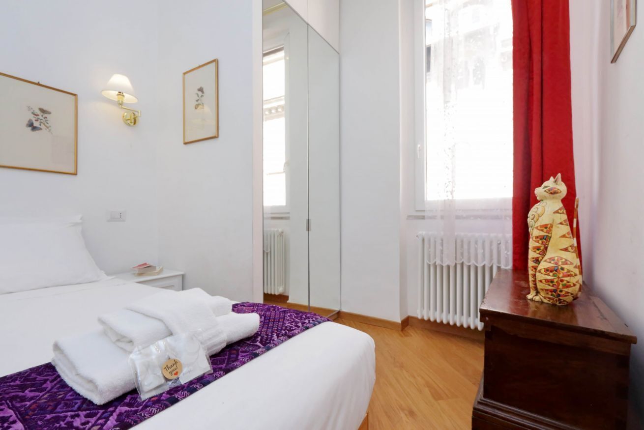 Una habitación de hotel con cama, mesita de noche, espejo, radiador y ventana con cortinas rojas.