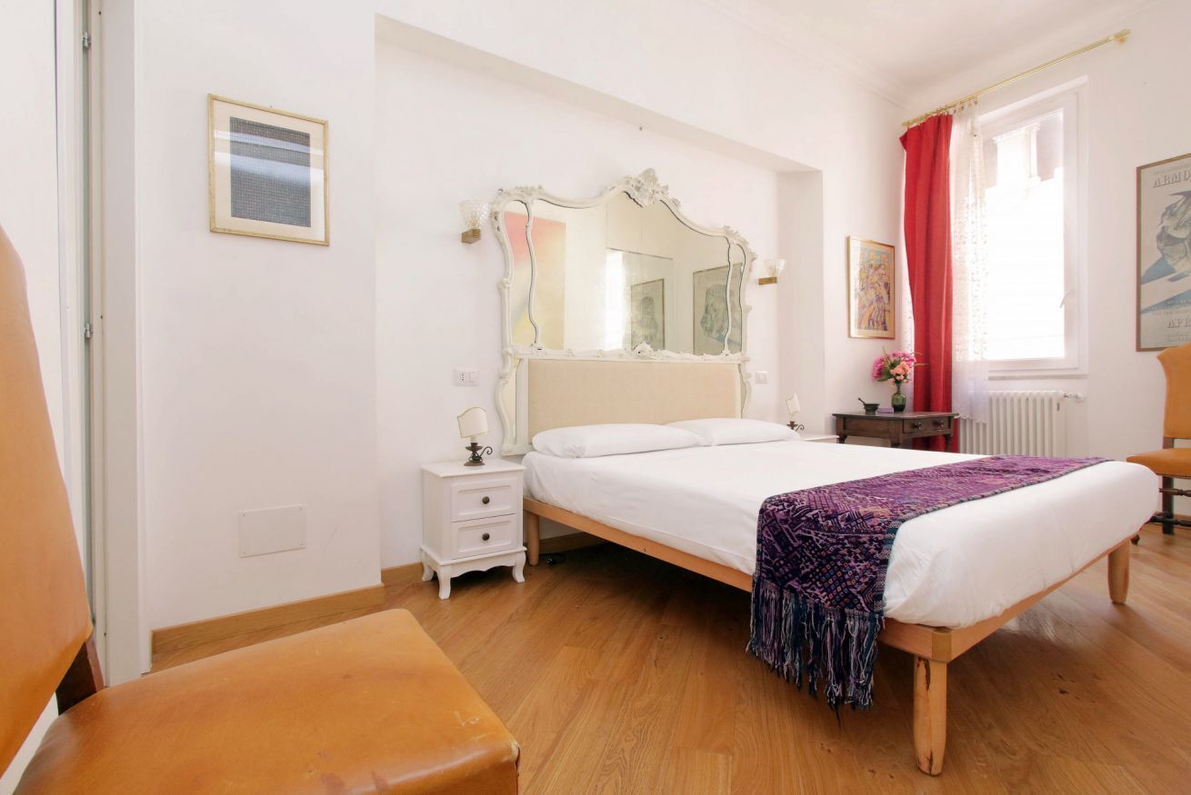 A bedroom with a bed, a nightstand, a chair, a mirror, and a window with curtains.