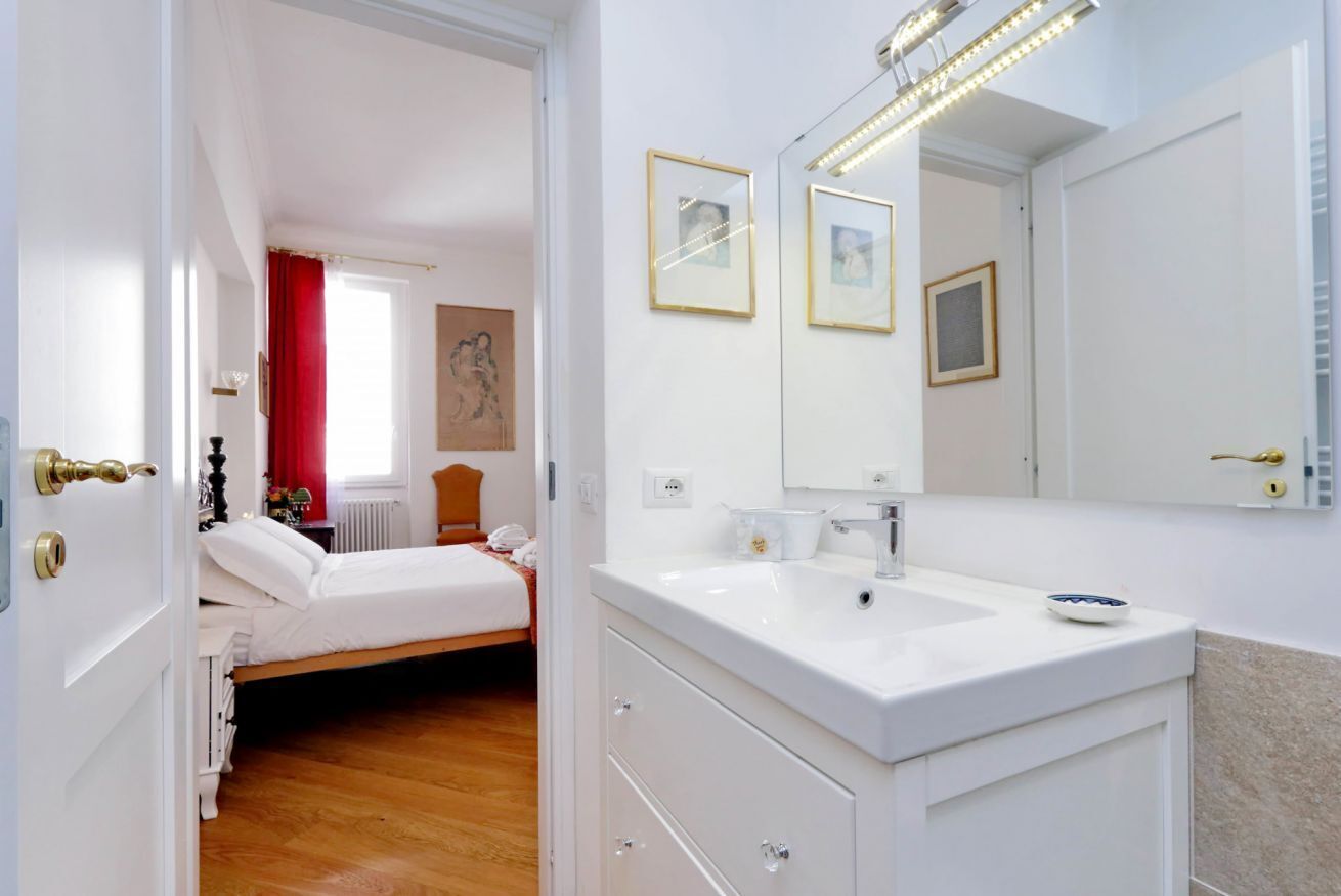 Una habitación de hotel con cama, un baño con lavamanos y espejo, y una puerta que da a otra habitación.