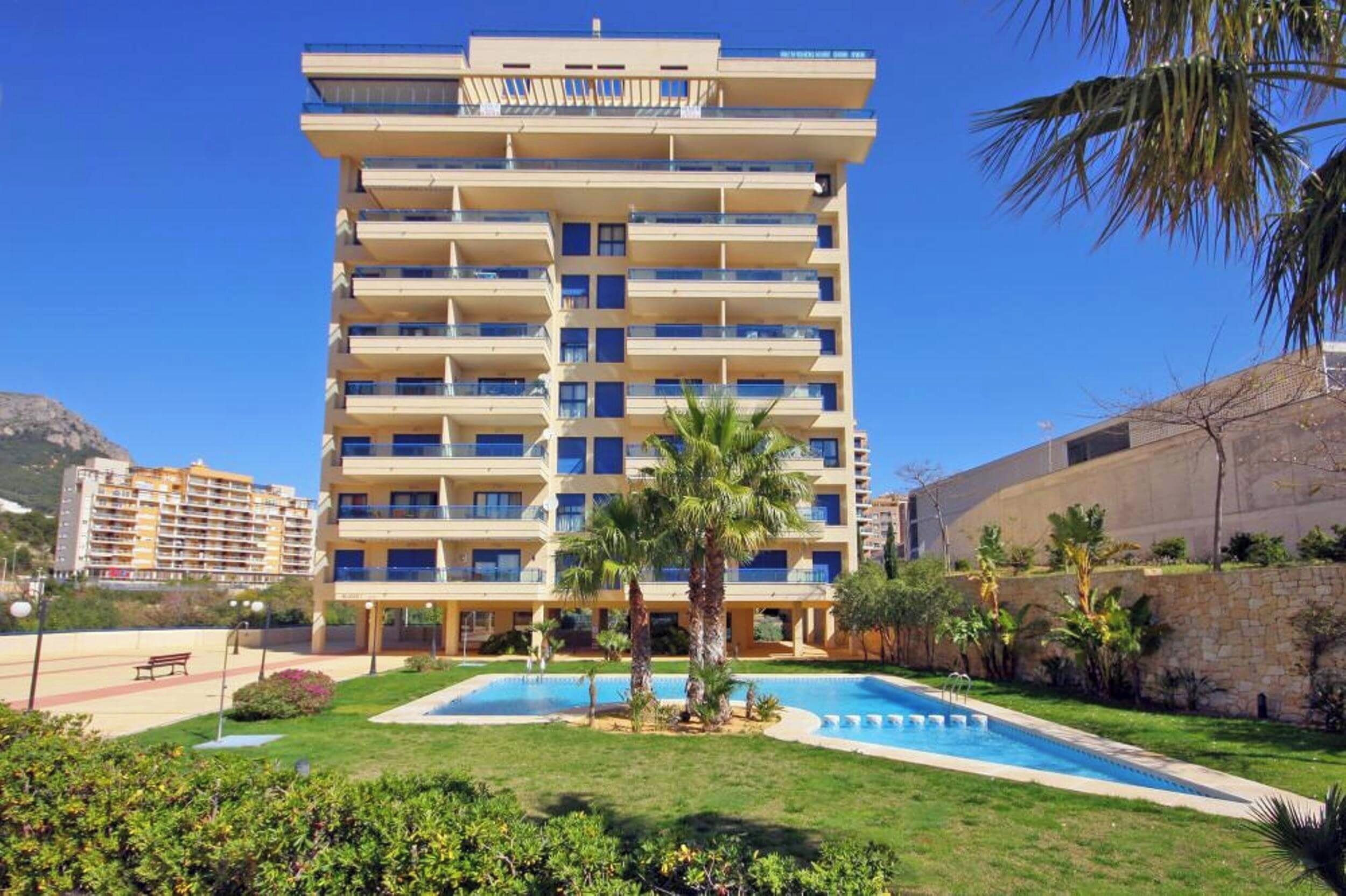 A multi-story hotel with a swimming pool, surrounded by greenery and palm trees.
