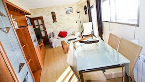Een compacte woonruimte met een bank, een glazen eettafel, rieten stoelen, een houten garderobe en een kleine kitchenette.