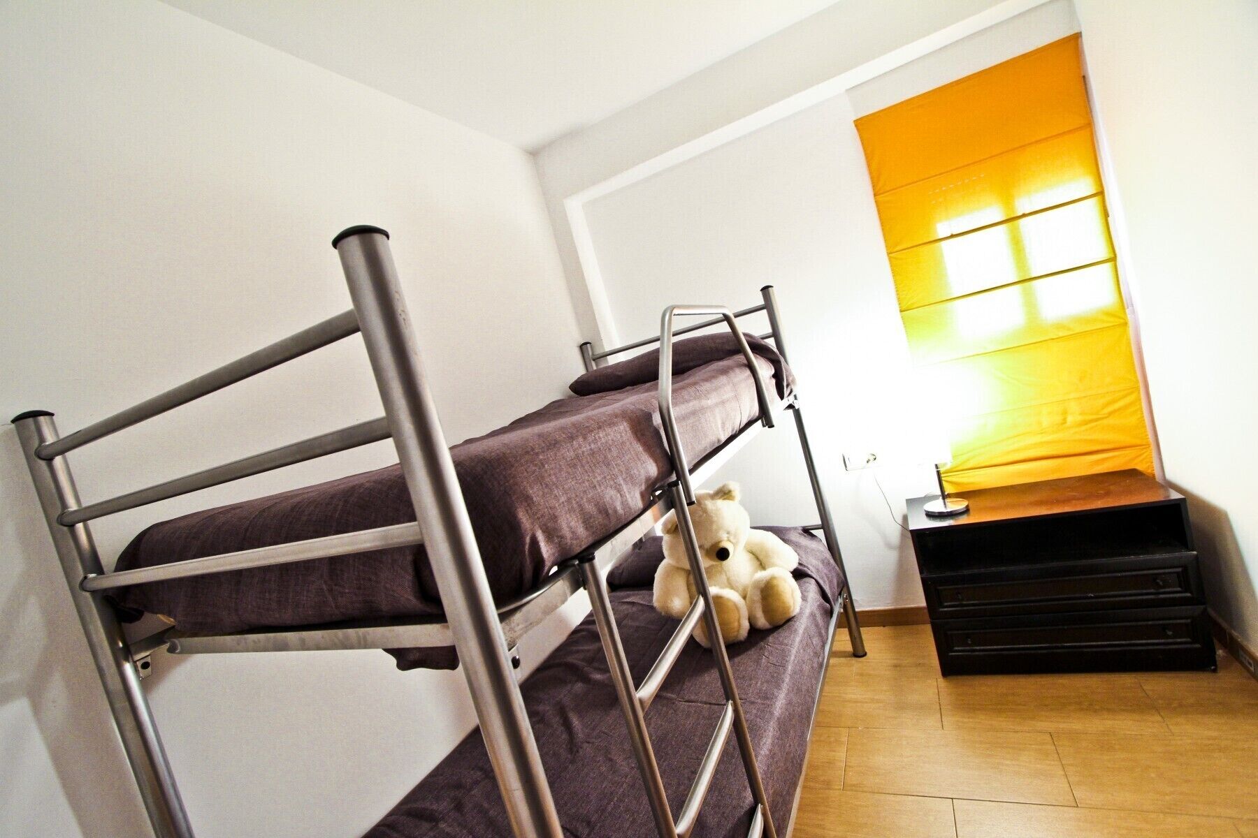 A bunk bed with a teddy bear, a small table, and a window with yellow blinds.