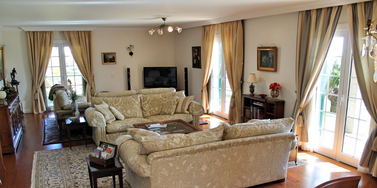 A spacious living room with a large beige sofa, a coffee table, and a television. There are curtains on the windows, and a chandelier hangs from the ceiling.
