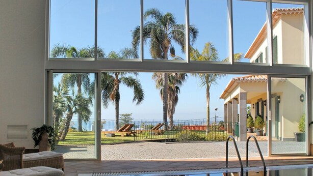 Una stanza con ampie finestre che si affaccia su una zona piscina con palme e lettini.