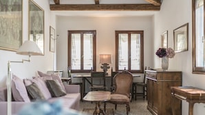 Una habitación acogedora con un sofá, una silla de madera, una mesita y un armario. Hay dos ventanas con cortinas, una lámpara sobre la mesa y cuadros enmarcados en las paredes.