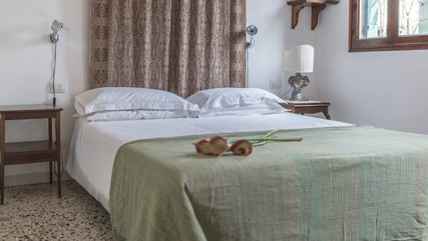 Una cama bien hecha con sábanas blancas, un edredón verde y un par de flores sobre la cama. Se ve una mesita de noche con una lámpara y un estante de pared con un objeto decorativo.